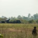 The image shows a military field exercise or deployment in a tropical or subtropical open terrain environment. A large truck-mounted multiple launch rocket system (MLRS) is positioned in the middle distance, with a soldier in camouflage uniform visible in the foreground. The setting appears to be a military training ground or operational area bordered by dense treeline.