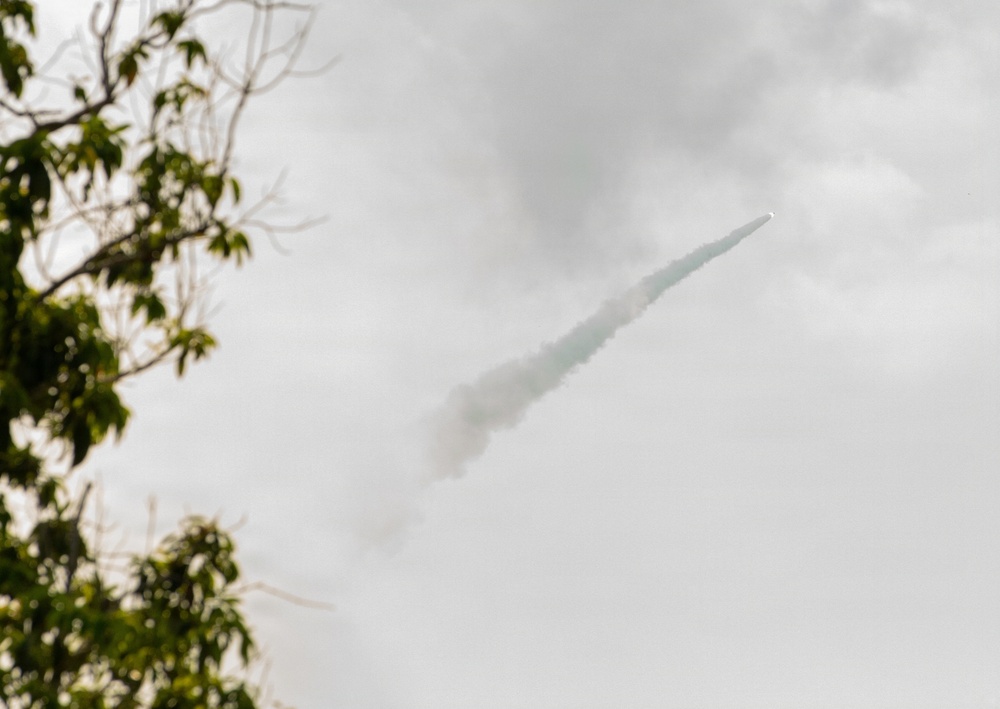 The image shows a rocket or missile in flight against an overcast sky, leaving a distinct white smoke trail behind it. The projectile is traveling at an upward diagonal angle from lower-left to upper-right. Green tree branches are visible in the blurred foreground on the left side of the frame.