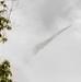 The image shows a rocket or missile in flight against an overcast sky, leaving a distinct white smoke trail behind it. The projectile is traveling at an upward diagonal angle from lower-left to upper-right. Green tree branches are visible in the blurred foreground on the left side of the frame.