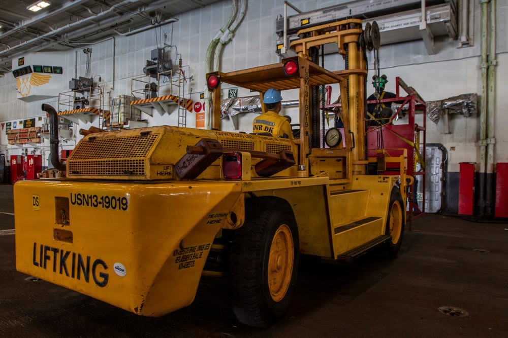 A large yellow industrial forklift branded 'LIFTKING' with the identifier 'USN13-19019' is being operated inside what appears to be a U.S. Navy ship's hangar bay. A single operator wearing a blue hard hat and yellow vest is seated at the controls. The interior space features white-painted walls, overhead piping, safety signage, and various equipment typical of a naval vessel's below-deck workspace.