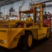 A large yellow industrial forklift branded 'LIFTKING' with the identifier 'USN13-19019' is being operated inside what appears to be a U.S. Navy ship's hangar bay. A single operator wearing a blue hard hat and yellow vest is seated at the controls. The interior space features white-painted walls, overhead piping, safety signage, and various equipment typical of a naval vessel's below-deck workspace.