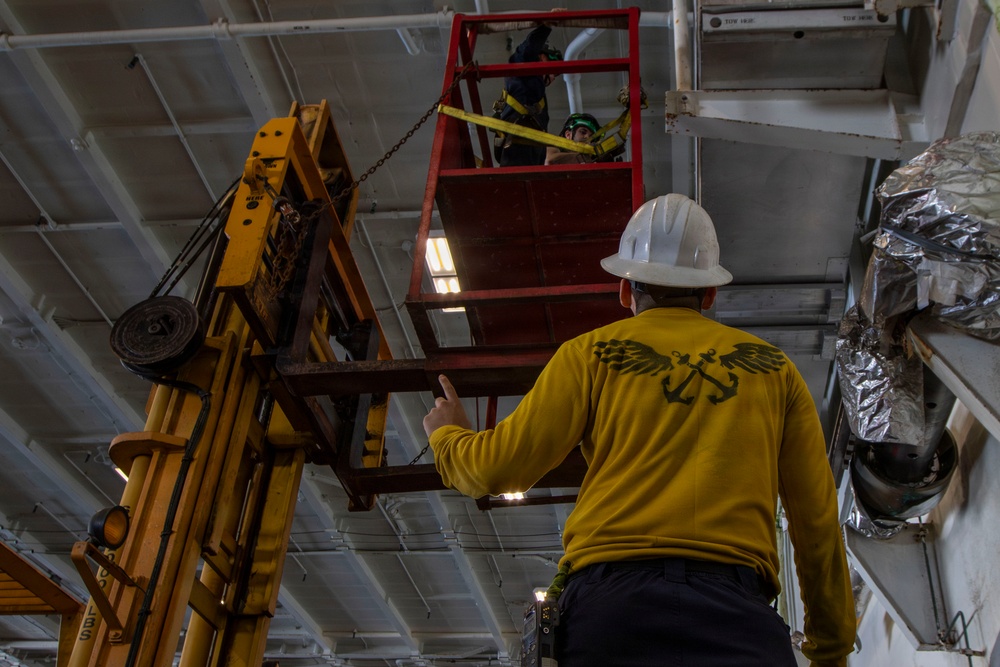 The image shows workers performing maintenance or installation work at height inside what appears to be a large industrial or naval vessel interior. A worker in a yellow long-sleeve shirt with an eagle-and-anchor graphic and white hard hat is directing operations from below, while at least one other worker is positioned in an elevated red personnel platform. A yellow forklift or material handler is being used to support or lift equipment near the ceiling.