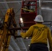 The image shows workers performing maintenance or installation work at height inside what appears to be a large industrial or naval vessel interior. A worker in a yellow long-sleeve shirt with an eagle-and-anchor graphic and white hard hat is directing operations from below, while at least one other worker is positioned in an elevated red personnel platform. A yellow forklift or material handler is being used to support or lift equipment near the ceiling.