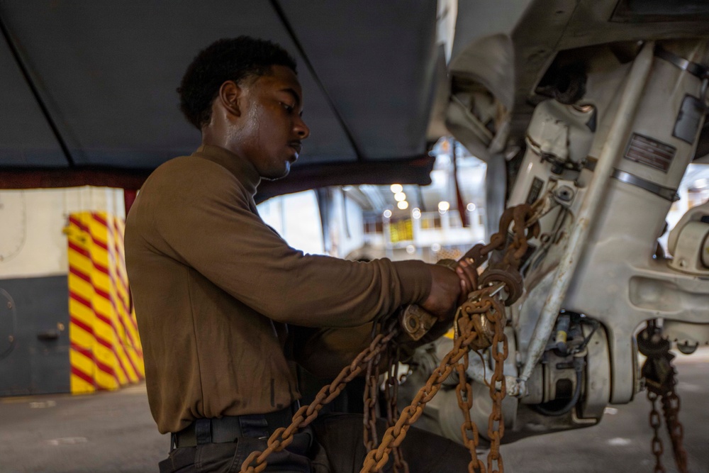 A young male service member in a brown military uniform is working with heavy rust-colored chains attached to what appears to be military aircraft landing gear or undercarriage equipment. The work is taking place in what appears to be a hangar bay, likely aboard a naval vessel based on the visible yellow and red hazard-striped structural elements in the background. The individual appears focused on securing or releasing chain tie-downs.