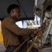 A young male service member in a brown military uniform is working with heavy rust-colored chains attached to what appears to be military aircraft landing gear or undercarriage equipment. The work is taking place in what appears to be a hangar bay, likely aboard a naval vessel based on the visible yellow and red hazard-striped structural elements in the background. The individual appears focused on securing or releasing chain tie-downs.