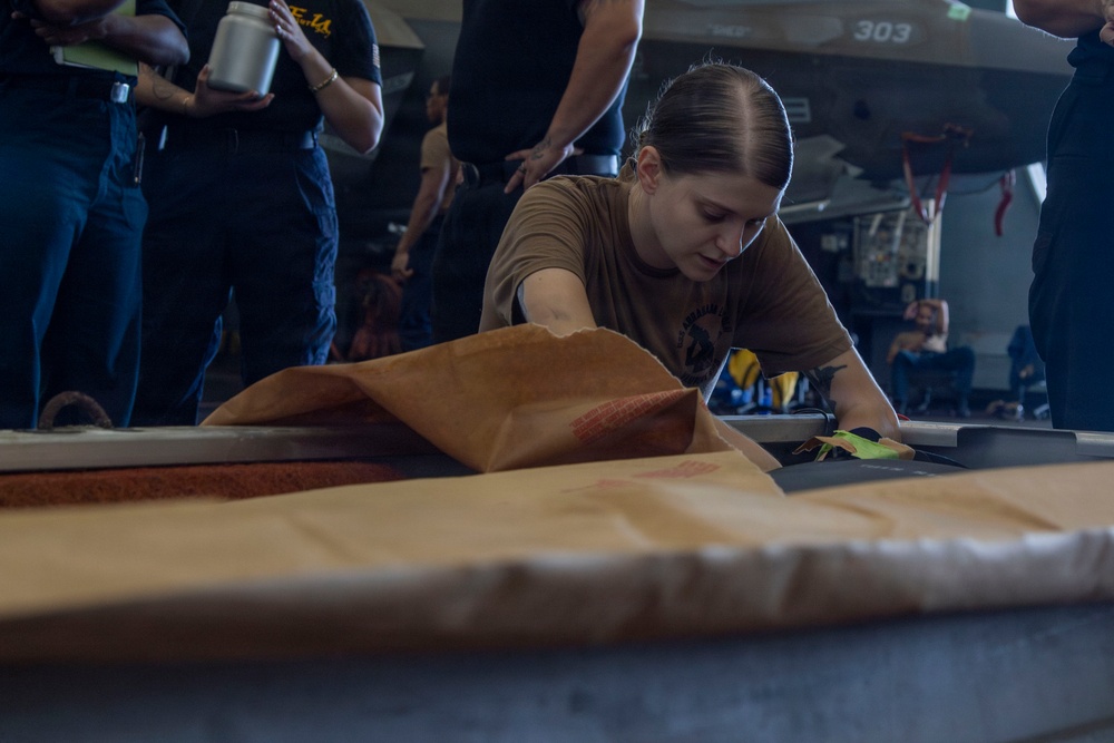 A person wearing a brown/tan military-style t-shirt is working at a table in what appears to be an aircraft hangar bay, handling brown paper or material. In the background, a military aircraft marked '303' is visible, along with several other personnel observing or standing nearby. The setting strongly suggests a naval vessel hangar deck environment.