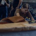 A person wearing a brown/tan military-style t-shirt is working at a table in what appears to be an aircraft hangar bay, handling brown paper or material. In the background, a military aircraft marked '303' is visible, along with several other personnel observing or standing nearby. The setting strongly suggests a naval vessel hangar deck environment.