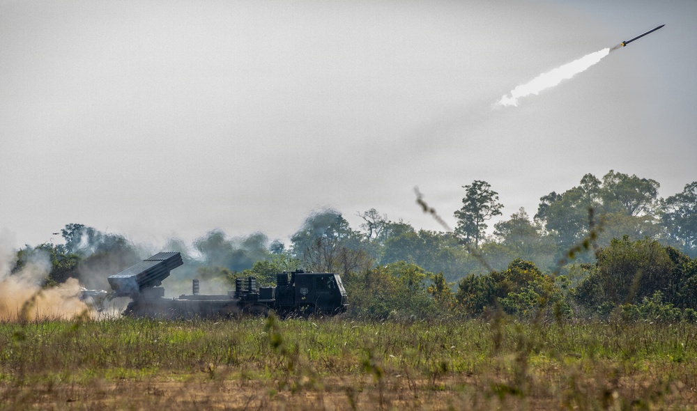 A military multiple launch rocket system (MLRS) mounted on a truck chassis is actively firing rockets in an open field setting. At least one rocket is clearly visible in flight, trailing a white smoke plume against an overcast sky. The scene appears to be a live-fire military exercise or demonstration, with significant smoke and dust surrounding the launcher.