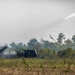 A military multiple launch rocket system (MLRS) mounted on a truck chassis is actively firing rockets in an open field setting. At least one rocket is clearly visible in flight, trailing a white smoke plume against an overcast sky. The scene appears to be a live-fire military exercise or demonstration, with significant smoke and dust surrounding the launcher.