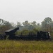 The image shows a military multiple launch rocket system (MLRS) mounted on a heavy truck, positioned in an open grassy field with dense tropical vegetation in the background. The launcher pod is elevated at an angle, suggesting a firing or ready position. At least one uniformed personnel is visible near the vehicle.