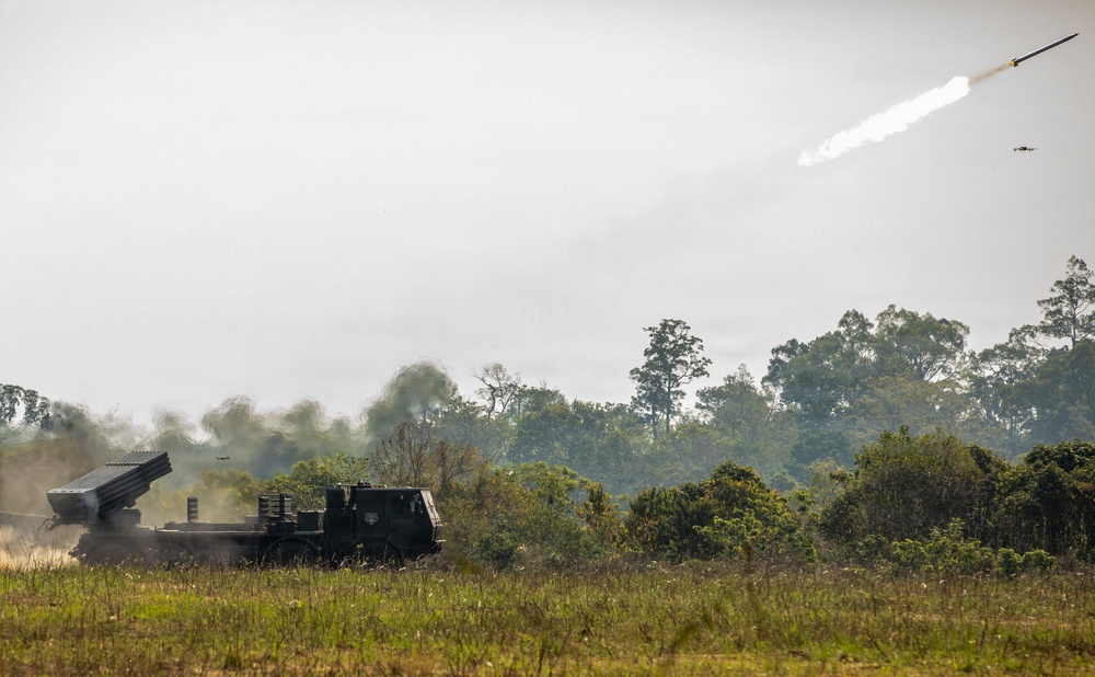 A multiple launch rocket system (MLRS) mounted on a military vehicle is firing a rocket in an open field environment. A rocket with a visible exhaust trail is captured mid-flight ascending into an overcast sky. A small drone or aircraft is also visible in the upper right portion of the image.