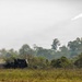 A multiple launch rocket system (MLRS) mounted on a military vehicle is firing a rocket in an open field environment. A rocket with a visible exhaust trail is captured mid-flight ascending into an overcast sky. A small drone or aircraft is also visible in the upper right portion of the image.