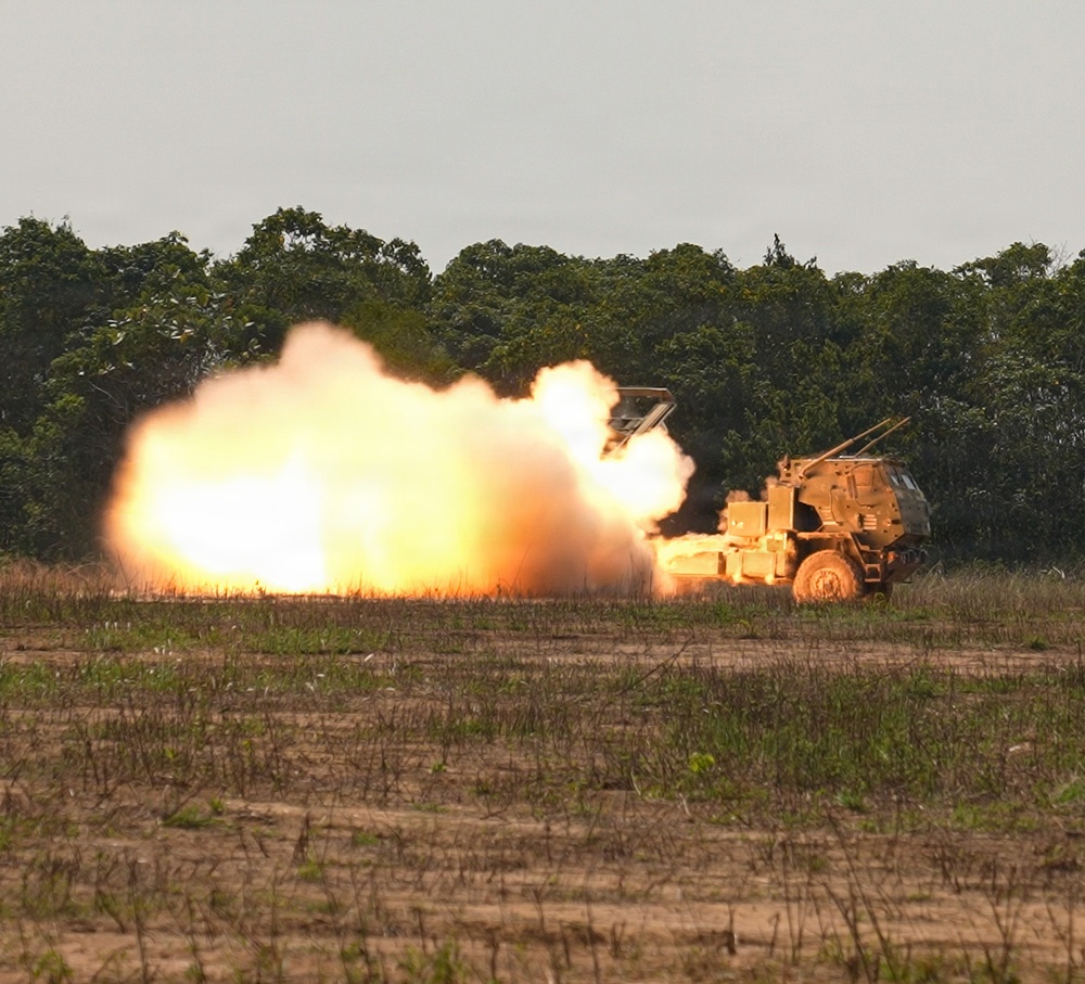 A wheeled military rocket artillery system is captured mid-launch, producing a large fireball and smoke cloud to its left side. The vehicle appears to be a HIMARS or similar multiple launch rocket system (MLRS) in a desert tan/sand color scheme. The launch is taking place on an open field with treeline visible in the background.