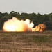 A wheeled military rocket artillery system is captured mid-launch, producing a large fireball and smoke cloud to its left side. The vehicle appears to be a HIMARS or similar multiple launch rocket system (MLRS) in a desert tan/sand color scheme. The launch is taking place on an open field with treeline visible in the background.