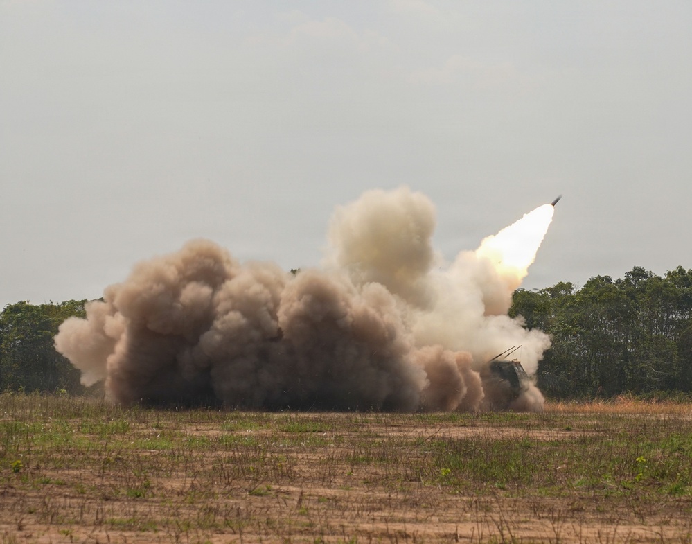 A military rocket artillery system is captured mid-launch, with a rocket visibly ascending at an angle in the upper right portion of the frame. A large billowing cloud of brown and white smoke surrounds the launch vehicle, which is partially visible on the right side. The scene takes place on an open grassy field with a treeline in the background.