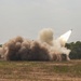 A military rocket artillery system is captured mid-launch, with a rocket visibly ascending at an angle in the upper right portion of the frame. A large billowing cloud of brown and white smoke surrounds the launch vehicle, which is partially visible on the right side. The scene takes place on an open grassy field with a treeline in the background.