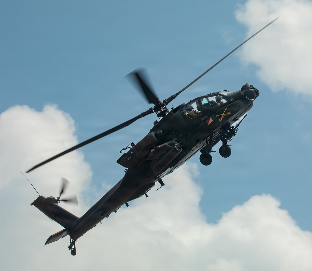 A military attack helicopter, identifiable as a Boeing AH-64 Apache, is photographed in flight against a partly cloudy blue sky. The helicopter is captured from a low angle, showing its underside and port side with rotor blades in motion. Two crew members are visible in the tandem cockpit, and the aircraft bears unit insignia on its fuselage.