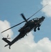A military attack helicopter, identifiable as a Boeing AH-64 Apache, is photographed in flight against a partly cloudy blue sky. The helicopter is captured from a low angle, showing its underside and port side with rotor blades in motion. Two crew members are visible in the tandem cockpit, and the aircraft bears unit insignia on its fuselage.
