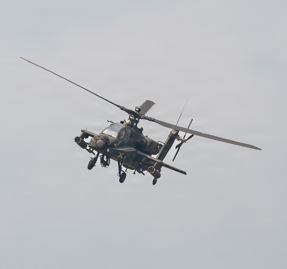 A military attack helicopter is photographed in flight against an overcast gray sky. The aircraft is viewed from a slightly below-front angle, revealing its weapons systems and tandem cockpit configuration. The helicopter appears to be in active flight with rotor blades in motion.
