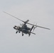A military attack helicopter is photographed in flight against an overcast gray sky. The aircraft is viewed from a slightly below-front angle, revealing its weapons systems and tandem cockpit configuration. The helicopter appears to be in active flight with rotor blades in motion.
