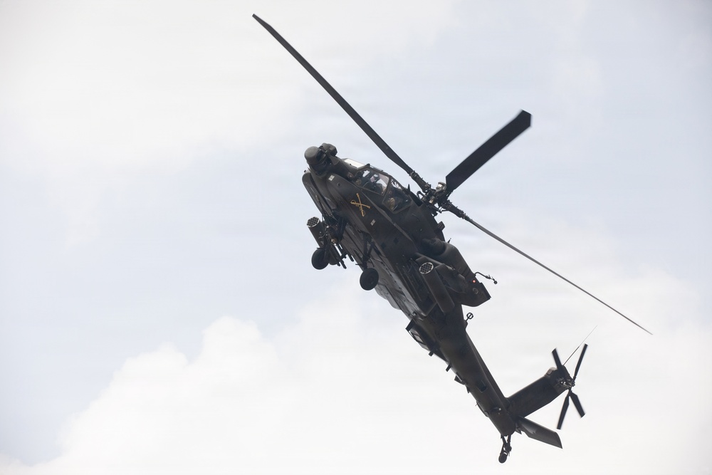 A military attack helicopter, consistent with an AH-64 Apache, is photographed from below at an angle against an overcast sky. The aircraft is in flight and appears to be banking or maneuvering, providing a clear view of its underside and rotor systems. A gold crossed-sabers insignia is visible on the fuselage, indicating U.S. Army Cavalry unit affiliation.