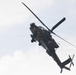 A military attack helicopter, consistent with an AH-64 Apache, is photographed from below at an angle against an overcast sky. The aircraft is in flight and appears to be banking or maneuvering, providing a clear view of its underside and rotor systems. A gold crossed-sabers insignia is visible on the fuselage, indicating U.S. Army Cavalry unit affiliation.