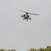 A military attack helicopter, consistent with an AH-64 Apache, is photographed in flight against an overcast gray sky. The helicopter is viewed from a slightly below-front angle, revealing its tandem cockpit, stub wings with weapon pylons, and sensor systems. Treetops are visible along the bottom edge of the frame.