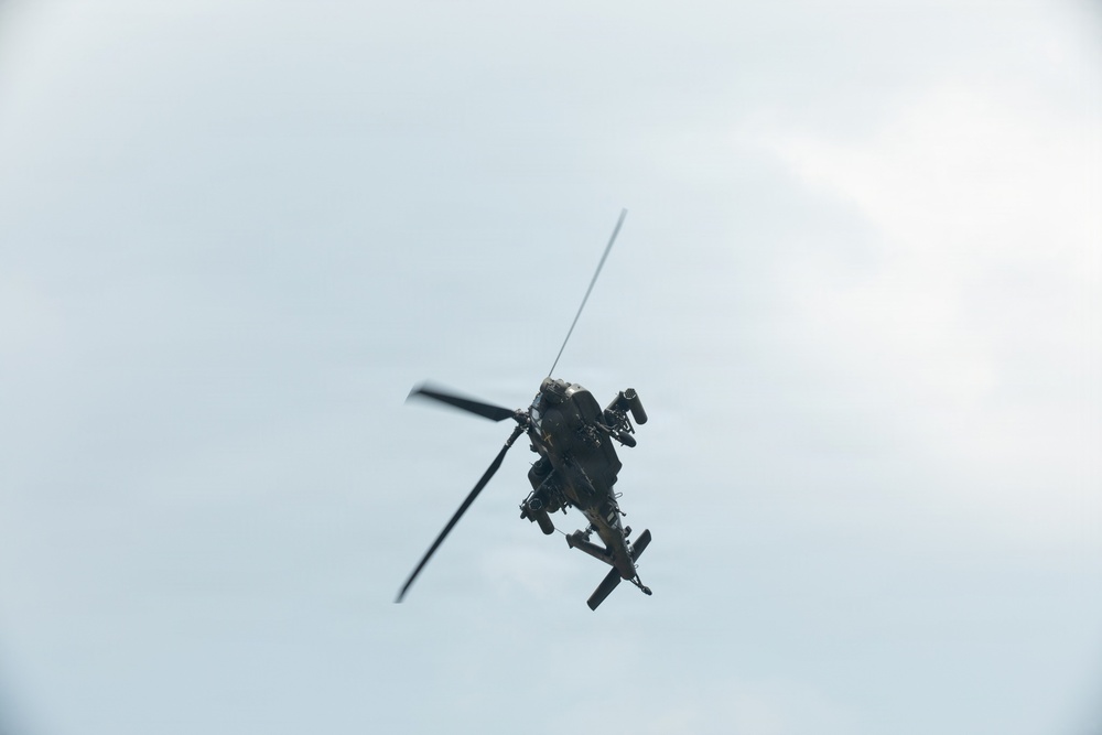 A military attack helicopter is photographed from below against an overcast sky. The aircraft appears to be in flight, viewed from a low angle showing its underside. The helicopter displays characteristics consistent with an attack helicopter design, featuring stub wings and weapons systems.