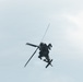 A military attack helicopter is photographed from below against an overcast sky. The aircraft appears to be in flight, viewed from a low angle showing its underside. The helicopter displays characteristics consistent with an attack helicopter design, featuring stub wings and weapons systems.
