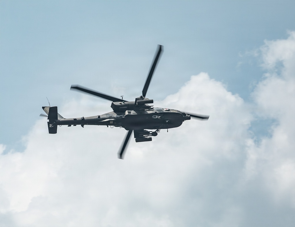 A military attack helicopter is photographed in flight against a partly cloudy sky. The aircraft appears to be an AH-64 Apache or similar attack helicopter, viewed from below at a slight angle. The rotor blades show motion blur, indicating active flight.