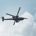 A military attack helicopter is photographed in flight against a partly cloudy sky. The aircraft appears to be an AH-64 Apache or similar attack helicopter, viewed from below at a slight angle. The rotor blades show motion blur, indicating active flight.