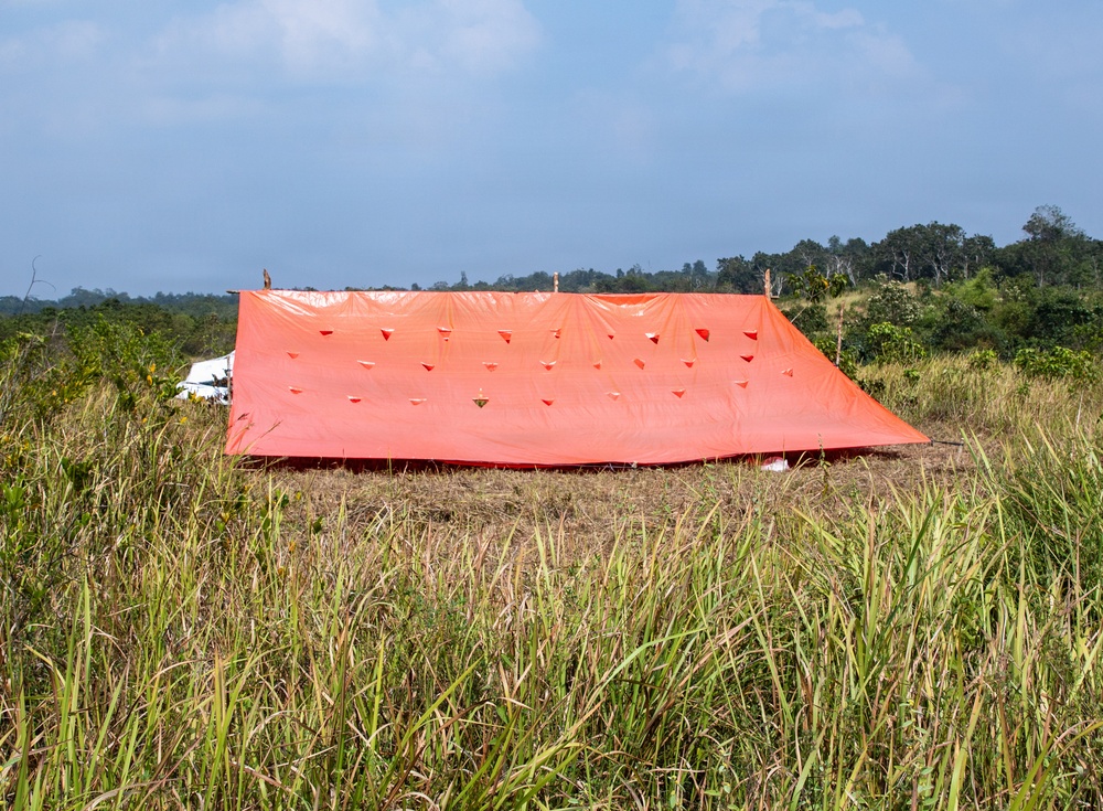 A large orange tarpaulin or emergency shelter cover is spread out flat over a cleared area of dry grass in what appears to be a tropical or subtropical field setting. The tarp is supported by several vertical poles and features multiple reinforced grommets or tie-down points across its surface. A white structure is partially visible in the background, surrounded by dense vegetation and trees.