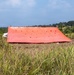 A large orange tarpaulin or emergency shelter cover is spread out flat over a cleared area of dry grass in what appears to be a tropical or subtropical field setting. The tarp is supported by several vertical poles and features multiple reinforced grommets or tie-down points across its surface. A white structure is partially visible in the background, surrounded by dense vegetation and trees.