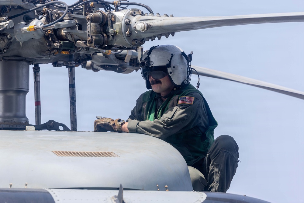 A U.S. military aviation crew member is performing maintenance or inspection work on top of a helicopter, positioned near the main rotor assembly. The individual is wearing a flight suit, aviation helmet with visor, and tactical gloves while crouching on the fuselage. The background suggests the helicopter may be operating over or near a body of water.
