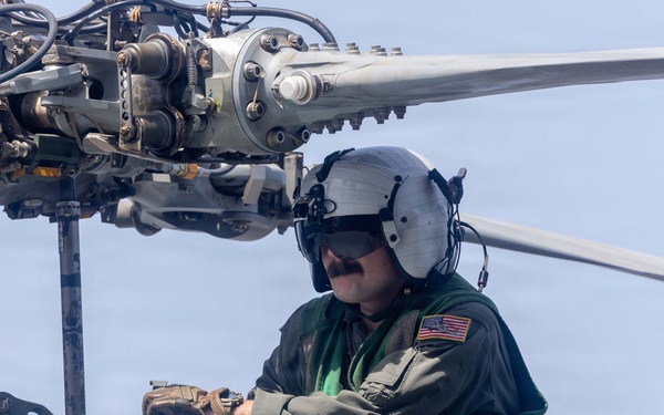 A U.S. military aviation crew member is performing maintenance or inspection work on top of a helicopter, positioned near the main rotor assembly. The individual is wearing a flight suit, aviation helmet with visor, and tactical gloves while crouching on the fuselage. The background suggests the helicopter may be operating over or near a body of water.