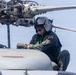 A U.S. military aviation crew member is performing maintenance or inspection work on top of a helicopter, positioned near the main rotor assembly. The individual is wearing a flight suit, aviation helmet with visor, and tactical gloves while crouching on the fuselage. The background suggests the helicopter may be operating over or near a body of water.