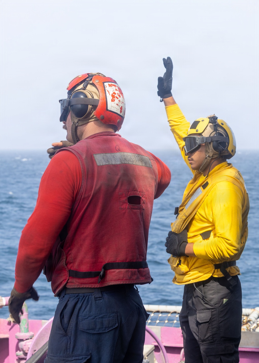 Two naval personnel are standing on what appears to be a ship's deck over open ocean. One individual wearing a red jersey/vest and red helmet with ear protection has their back to the camera, while a second individual in a yellow jersey and yellow helmet with goggles is raising one hand in a signaling gesture. Their color-coded gear is consistent with U.S. Navy flight deck crew roles.