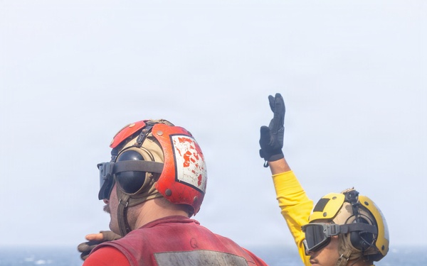 Two naval personnel are standing on what appears to be a ship's deck over open ocean. One individual wearing a red jersey/vest and red helmet with ear protection has their back to the camera, while a second individual in a yellow jersey and yellow helmet with goggles is raising one hand in a signaling gesture. Their color-coded gear is consistent with U.S. Navy flight deck crew roles.