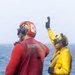 Two naval personnel are standing on what appears to be a ship's deck over open ocean. One individual wearing a red jersey/vest and red helmet with ear protection has their back to the camera, while a second individual in a yellow jersey and yellow helmet with goggles is raising one hand in a signaling gesture. Their color-coded gear is consistent with U.S. Navy flight deck crew roles.