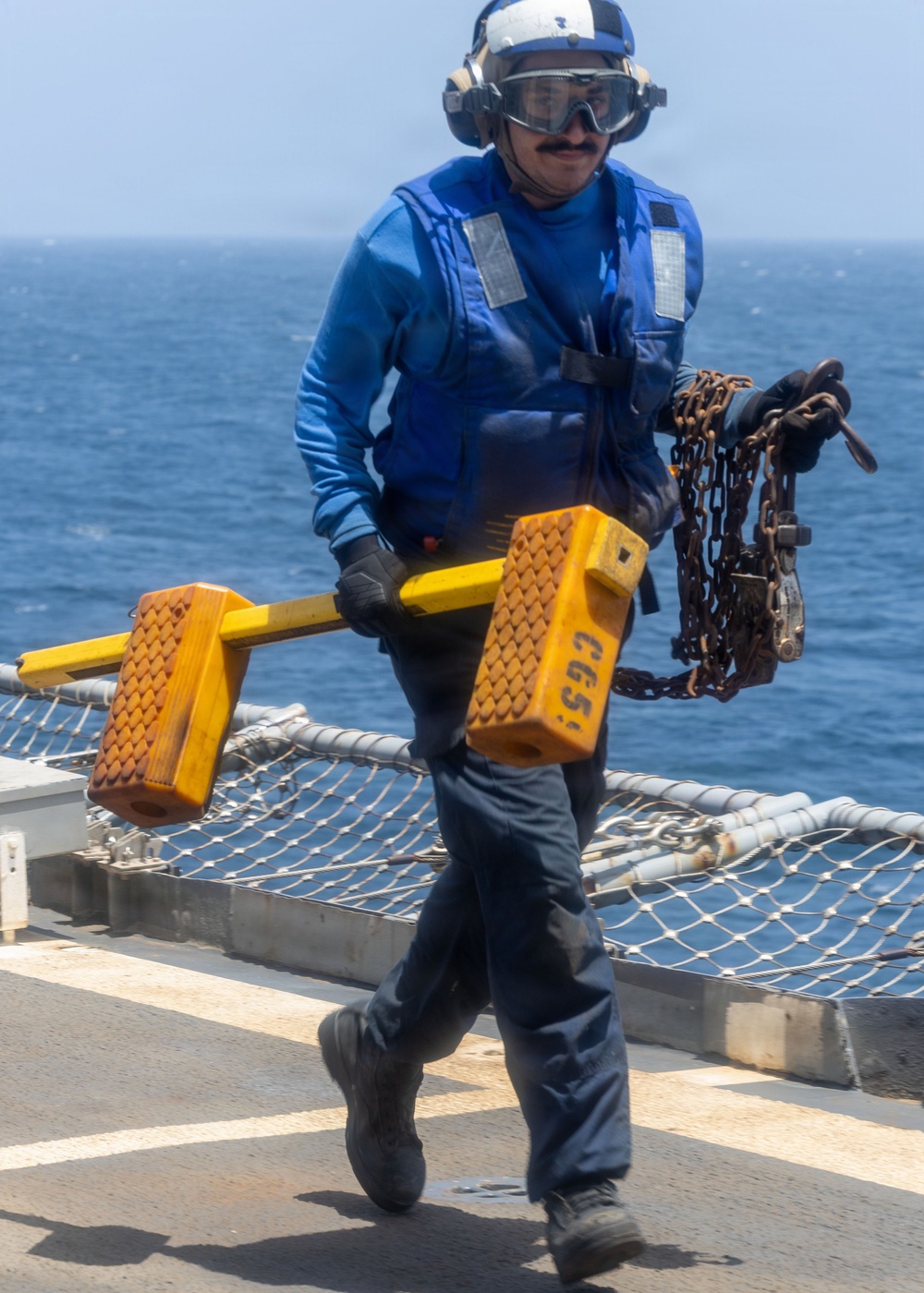 A naval aviation deck crew member dressed in blue flight deck gear is walking across what appears to be a ship's flight deck or helicopter landing pad. The sailor is carrying yellow wheel chocks marked 'CG65' and a heavy chain, standard equipment used to secure aircraft on naval vessels. The ocean is visible in the background, confirming an at-sea operational environment.