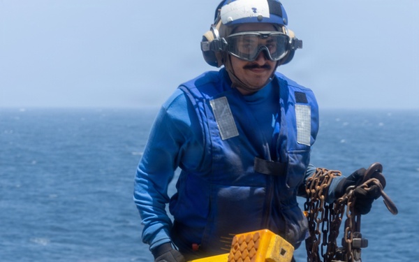 A naval aviation deck crew member dressed in blue flight deck gear is walking across what appears to be a ship's flight deck or helicopter landing pad. The sailor is carrying yellow wheel chocks marked 'CG65' and a heavy chain, standard equipment used to secure aircraft on naval vessels. The ocean is visible in the background, confirming an at-sea operational environment.