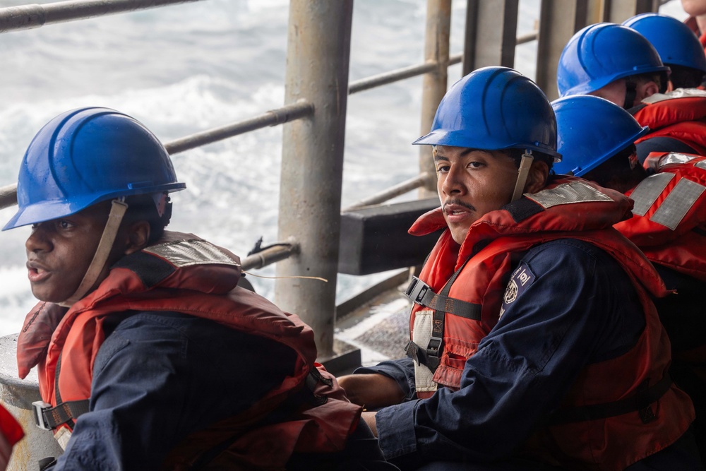 Several personnel wearing blue hard hats and orange life vests are seated on the deck of a vessel at sea. They appear to be engaged in a maritime operation or drill, with rough ocean water visible in the background. The individuals are dressed in navy blue uniforms beneath their safety equipment.