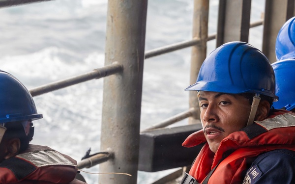 Several personnel wearing blue hard hats and orange life vests are seated on the deck of a vessel at sea. They appear to be engaged in a maritime operation or drill, with rough ocean water visible in the background. The individuals are dressed in navy blue uniforms beneath their safety equipment.
