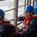 Several personnel wearing blue hard hats and orange life vests are seated on the deck of a vessel at sea. They appear to be engaged in a maritime operation or drill, with rough ocean water visible in the background. The individuals are dressed in navy blue uniforms beneath their safety equipment.