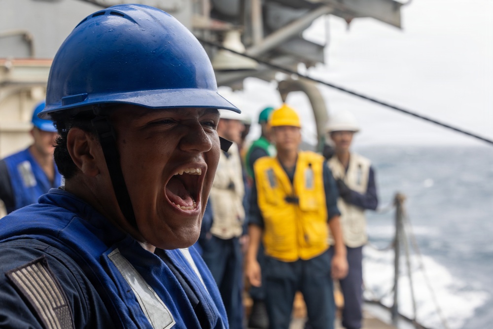A person wearing a blue hard hat and blue coveralls with a US flag patch is shouting or calling out loudly, likely issuing commands or relaying information. The scene takes place on the deck of a naval or military vessel at sea, with several other personnel visible in the background wearing various colored hard hats and safety vests. The setting and attire suggest a military or maritime operational exercise or working evolution.