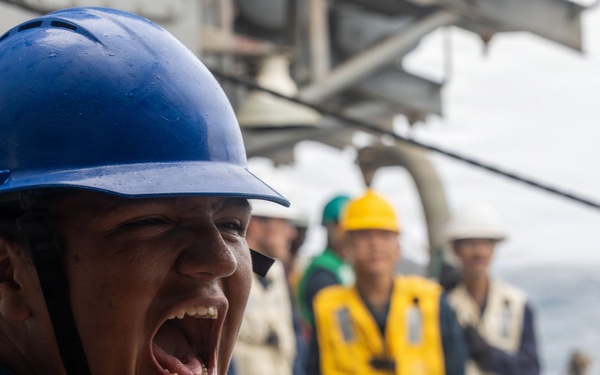 A person wearing a blue hard hat and blue coveralls with a US flag patch is shouting or calling out loudly, likely issuing commands or relaying information. The scene takes place on the deck of a naval or military vessel at sea, with several other personnel visible in the background wearing various colored hard hats and safety vests. The setting and attire suggest a military or maritime operational exercise or working evolution.