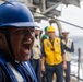 A person wearing a blue hard hat and blue coveralls with a US flag patch is shouting or calling out loudly, likely issuing commands or relaying information. The scene takes place on the deck of a naval or military vessel at sea, with several other personnel visible in the background wearing various colored hard hats and safety vests. The setting and attire suggest a military or maritime operational exercise or working evolution.