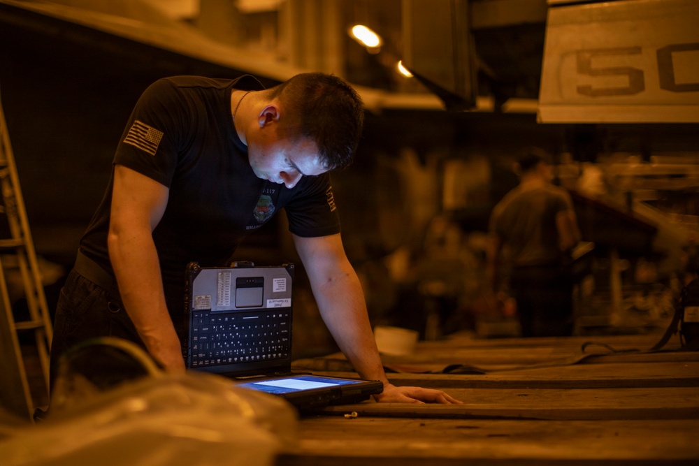 A male individual wearing a black military-style t-shirt with a U.S. flag patch on the sleeve is leaning over a surface and interacting with a ruggedized tablet/laptop device. The setting appears to be a dimly lit industrial or military hangar/bay environment with warm orange ambient lighting. The device he is using appears to be a military-grade ruggedized computing unit with a physical keyboard attachment.