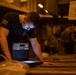 A male individual wearing a black military-style t-shirt with a U.S. flag patch on the sleeve is leaning over a surface and interacting with a ruggedized tablet/laptop device. The setting appears to be a dimly lit industrial or military hangar/bay environment with warm orange ambient lighting. The device he is using appears to be a military-grade ruggedized computing unit with a physical keyboard attachment.
