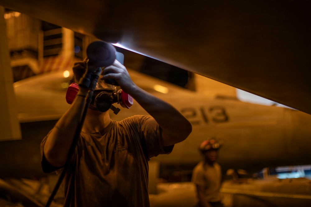 A maintenance technician is inspecting the underside of a military aircraft using a handheld flashlight in a low-light environment, likely inside an aircraft carrier hangar bay. The aircraft bears the number '213' on its fuselage. A second crew member wearing a helmet is visible in the background.